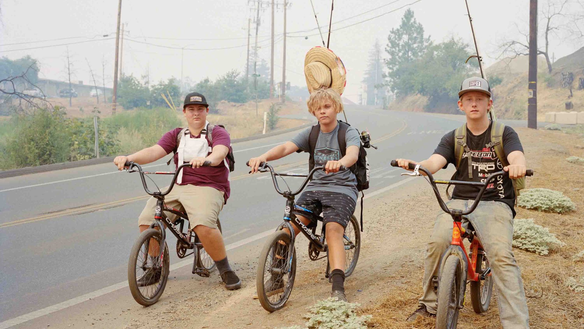 Three boys on bikes with fishing gear on a smoky road.
